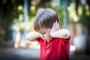 A 4 year old autistic child in a red shirt closing his ears with hands as if protecting from noise. Autism concept, Asperger syndrome, loud noise, scared little kid, parents divorce trauma.